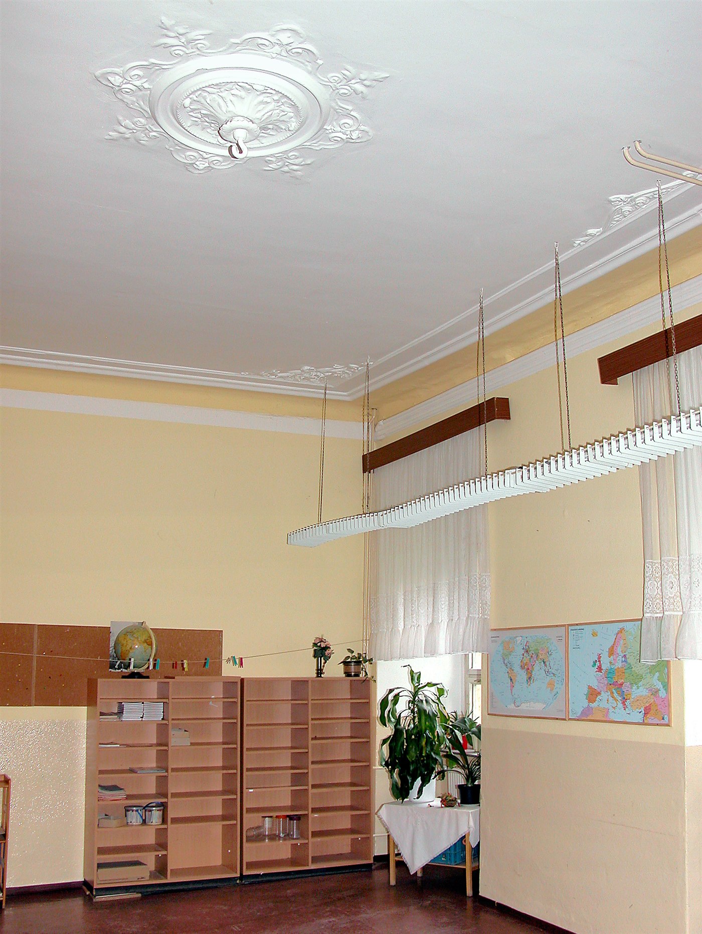 Wysoka sala szkolna ze sztukaterią na suficie, regałami z globusem i mapami na ścianie, bez osób.